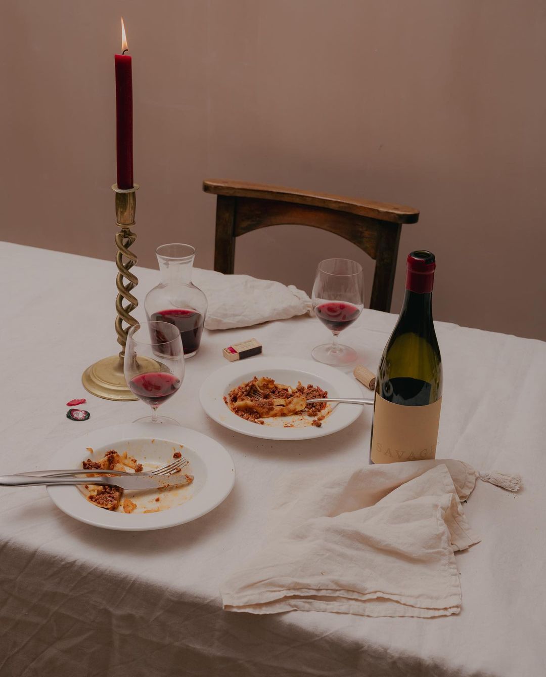 Still life of a table setting with plates of pasta, wine glasses filled with red wine, a candle, and a wine bottle, set indoors.