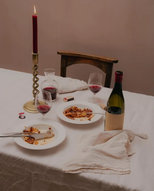 Still life of a table setting with plates of pasta, wine glasses filled with red wine, a candle, and a wine bottle, set indoors.
