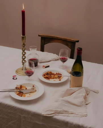 Still life of a table setting with plates of pasta, wine glasses filled with red wine, a candle, and a wine bottle, set indoors.