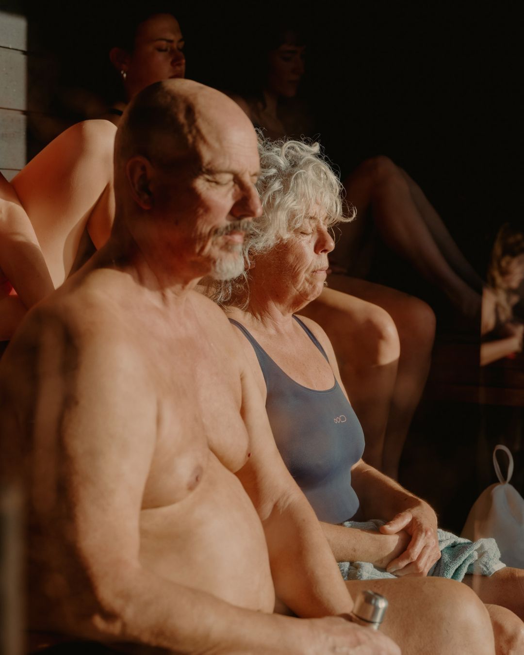 Several adults relax indoors in a naturally lit sauna. An older bald man and gray-haired woman are in the foreground.