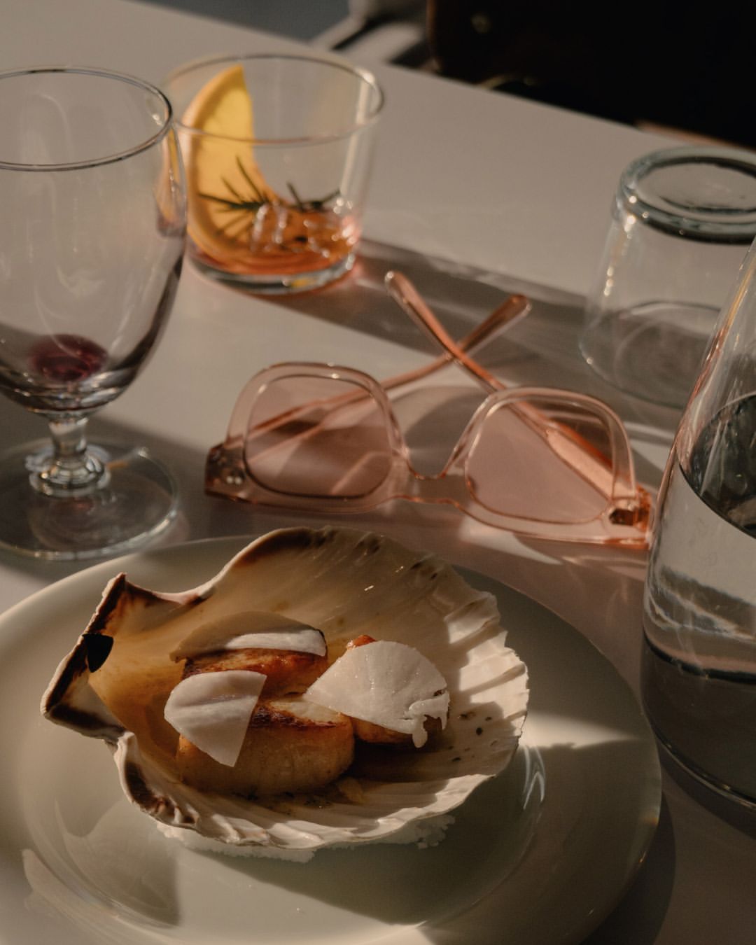 Seared scallops with white slices in seashell plate with stemware, water bottle, pink eyeglasses and orange slice drink on a white table.