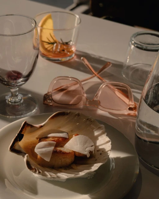 Seared scallops with white slices in seashell plate with stemware, water bottle, pink eyeglasses and orange slice drink on a white table.