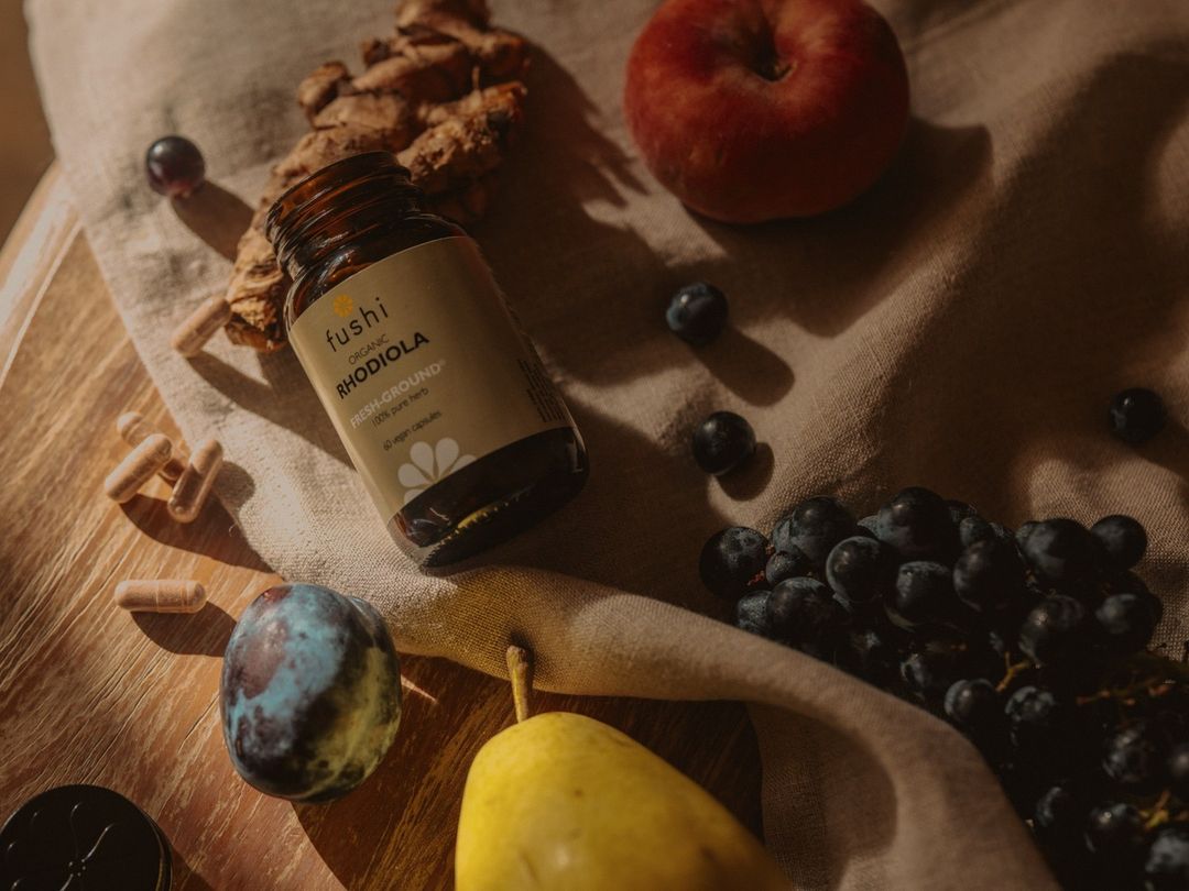 Still life of a brown supplement bottle of Rhodiola capsules surrounded by an assortment of fresh fruit on a rustic linen surface.