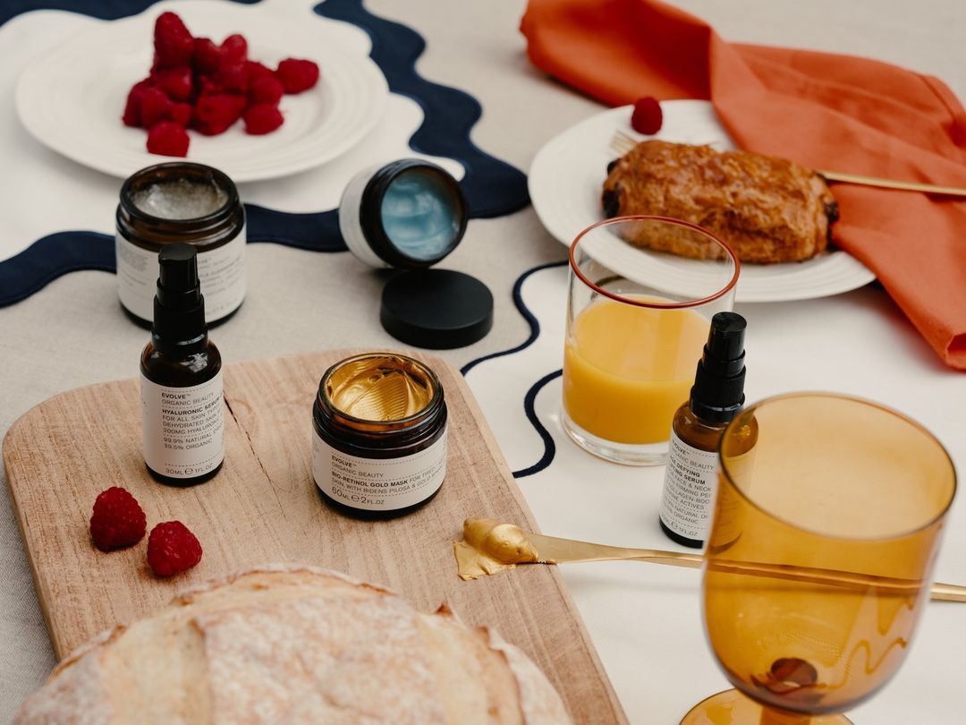 Still life of Evolve Organic Beauty skincare products, bread, pastries, juice, and raspberries arranged on a table with a patterned tablecloth.