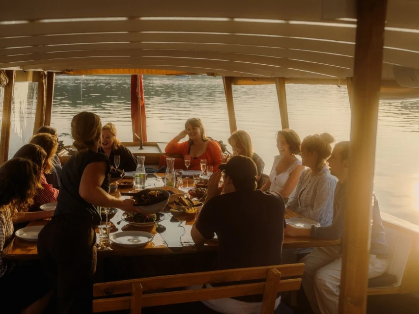 A group of people are sitting around a table on a boat, enjoying a meal with drinks, surrounded by calm waters in golden lighting.