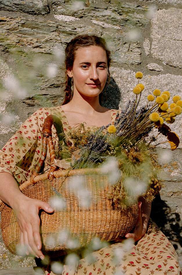 Young woman in a floral dress holding a wicker basket filled with dried flowers stands in front of a stone wall on a sunny day.