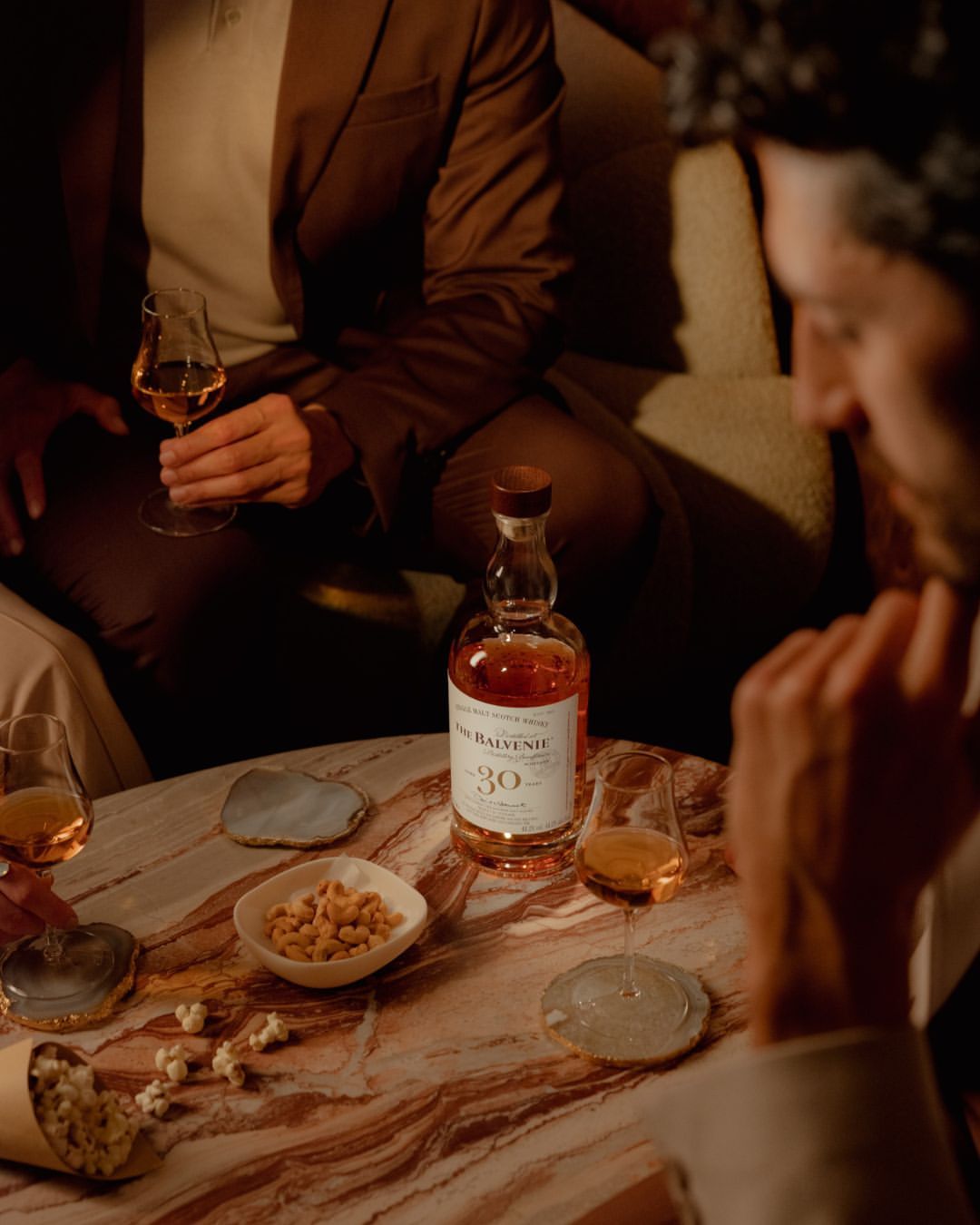 A bottle of Balvenie 30 whiskey sits on a marble table among snacks and glasses, with three adults in suits, in a moody indoor setting.