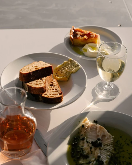 Still life of assorted food and drinks on white plates and glass containers, including sliced bread, tart, fish, and wine.