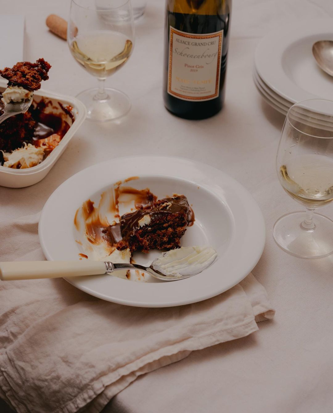 Overhead shot of a half-eaten chocolate dessert, white wine, and a wine bottle on a white tablecloth, suggesting a casual dining setting.