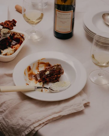 Overhead shot of a half-eaten chocolate dessert, white wine, and a wine bottle on a white tablecloth, suggesting a casual dining setting.