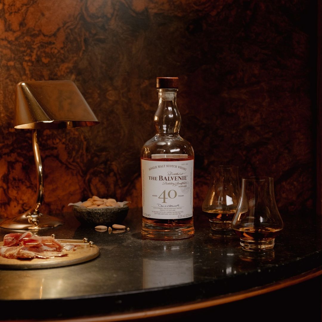 Still life shot of Balvenie Forty Single Malt Scotch Whisky bottle with two glasses, charcuterie board, almonds, and lamp on a table.