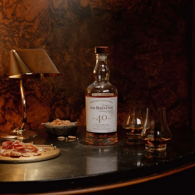 Still life shot of Balvenie Forty Single Malt Scotch Whisky bottle with two glasses, charcuterie board, almonds, and lamp on a table.