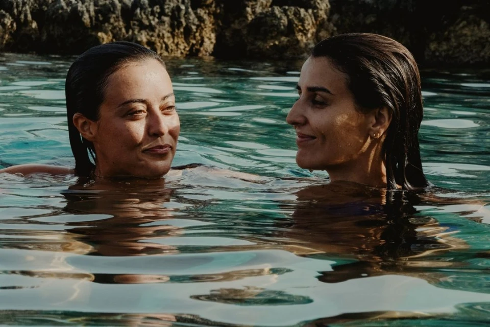 Two women are swimming in turquoise water on a sunny day, with rocky cliffs in the background. The women are looking at each other.