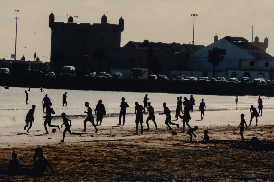 Silhouetted children playing soccer on a sandy beach against a backdrop of historic buildings and the soft light of late afternoon.