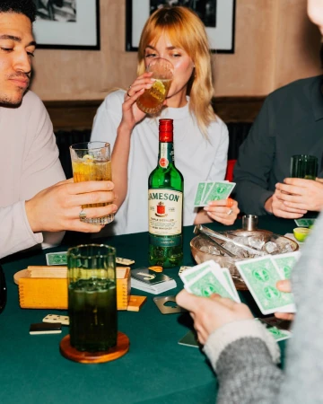 Several people are sitting around a green table with a bottle of Jameson, glasses of drinks, and playing cards in a casual setting.