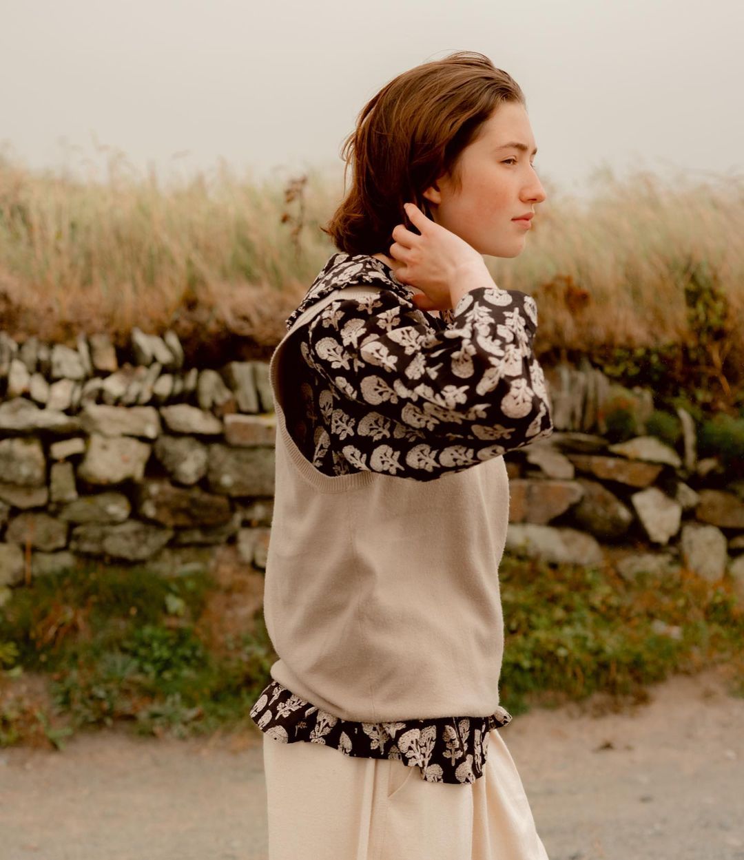 Young woman in skull-print top and apron stands outdoors with a stone wall and grassy field in the background, looking off to the side.
