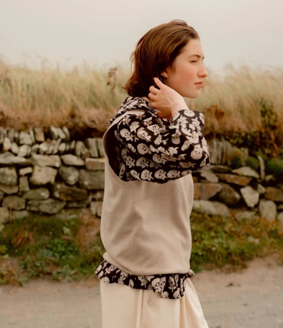 Young woman in skull-print top and apron stands outdoors with a stone wall and grassy field in the background, looking off to the side.