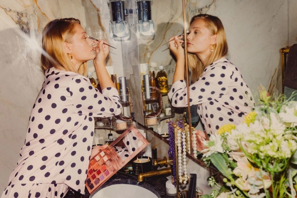 Young woman in polka-dot pajamas applies makeup in a marble-walled bathroom with flowers and toiletries visible in the mirror's reflection.