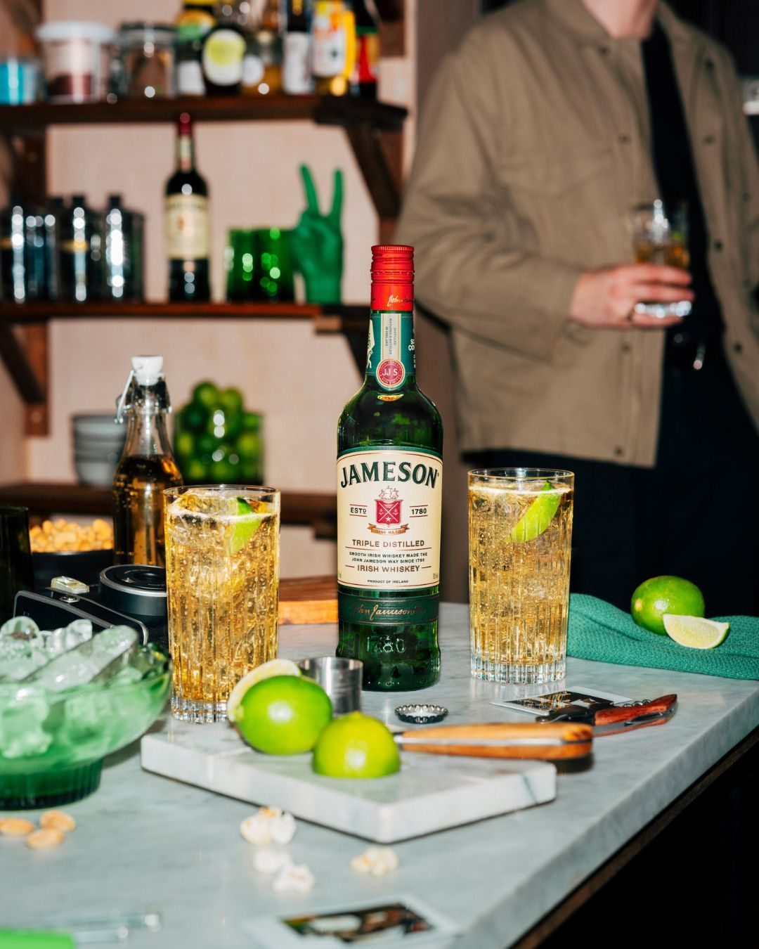 Bottle of Jameson whiskey surrounded by limes and cocktails on a marble countertop, featuring a person in the background holding a drink.