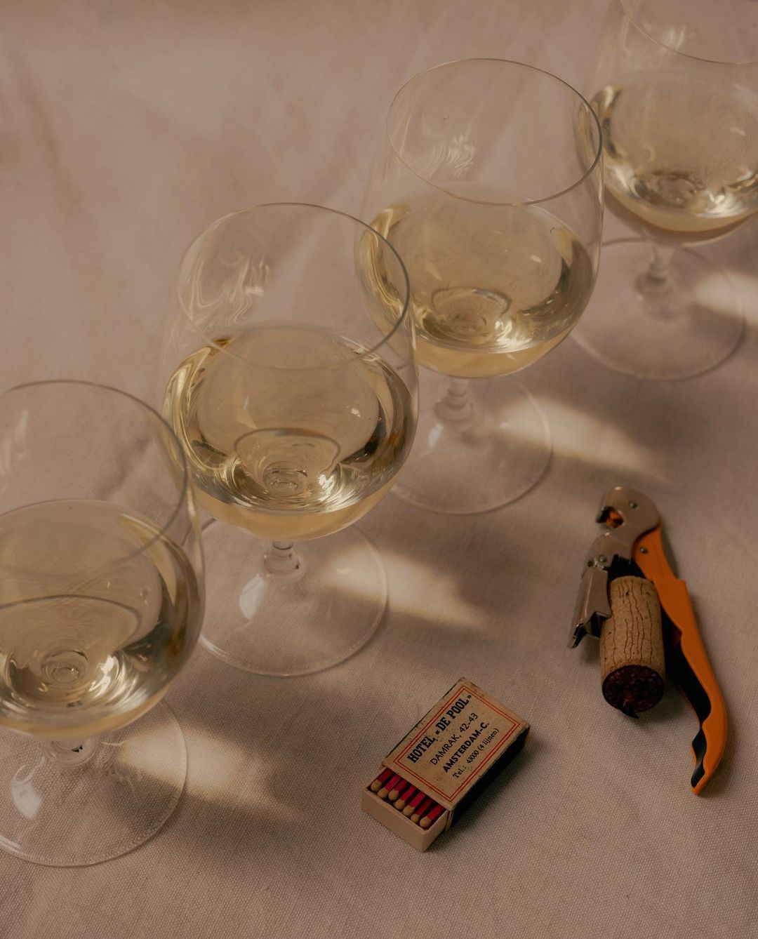 Still life of white wine in glasses, a corkscrew, and a matchbox on a white surface, creating a cozy and inviting atmosphere.