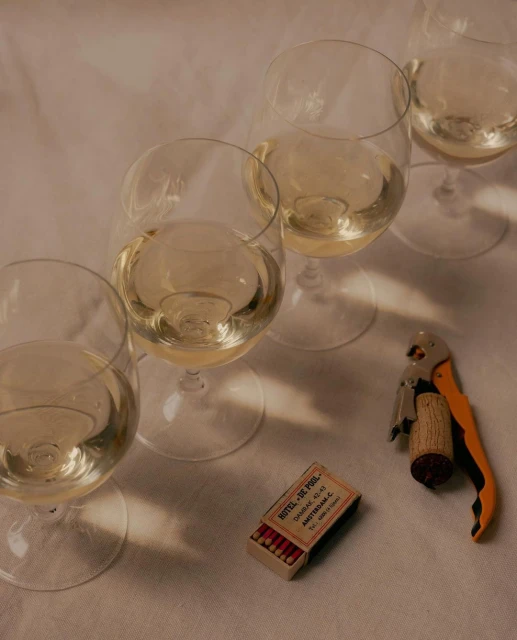 Still life of white wine in glasses, a corkscrew, and a matchbox on a white surface, creating a cozy and inviting atmosphere.