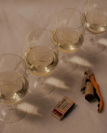 Still life of white wine in glasses, a corkscrew, and a matchbox on a white surface, creating a cozy and inviting atmosphere.