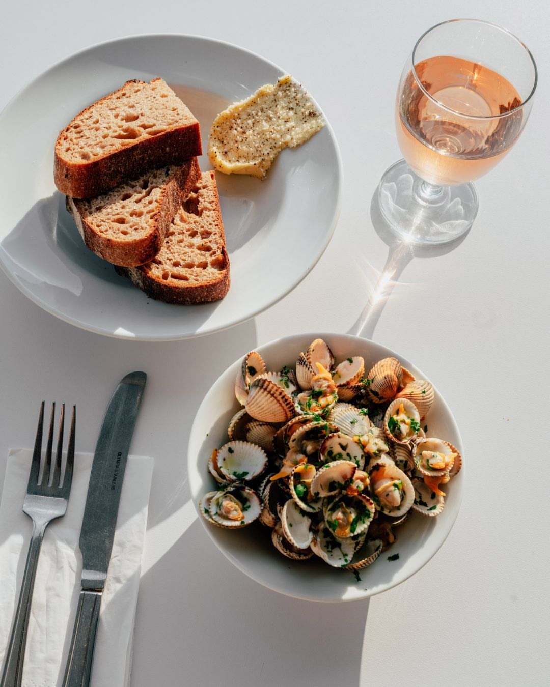 Overhead shot of a white table featuring a plate of sliced bread with butter, a bowl of cockles, and a glass of rose wine.
