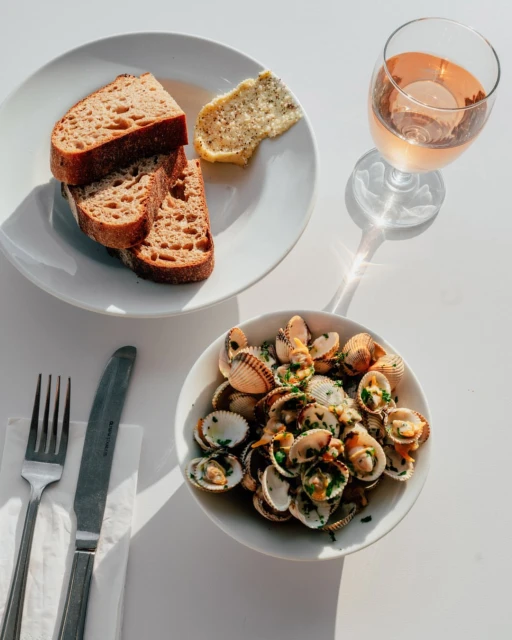Overhead shot of a white table featuring a plate of sliced bread with butter, a bowl of cockles, and a glass of rose wine.