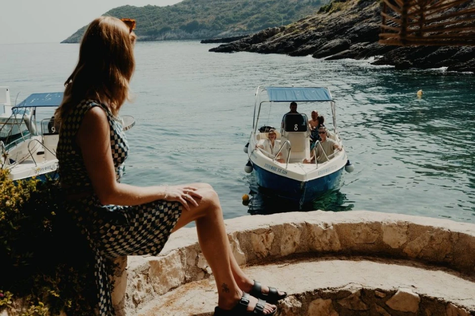Woman in a checkered dress sits on a stone ledge by the sea, looking at a motorboat with passengers near a rocky coastline.