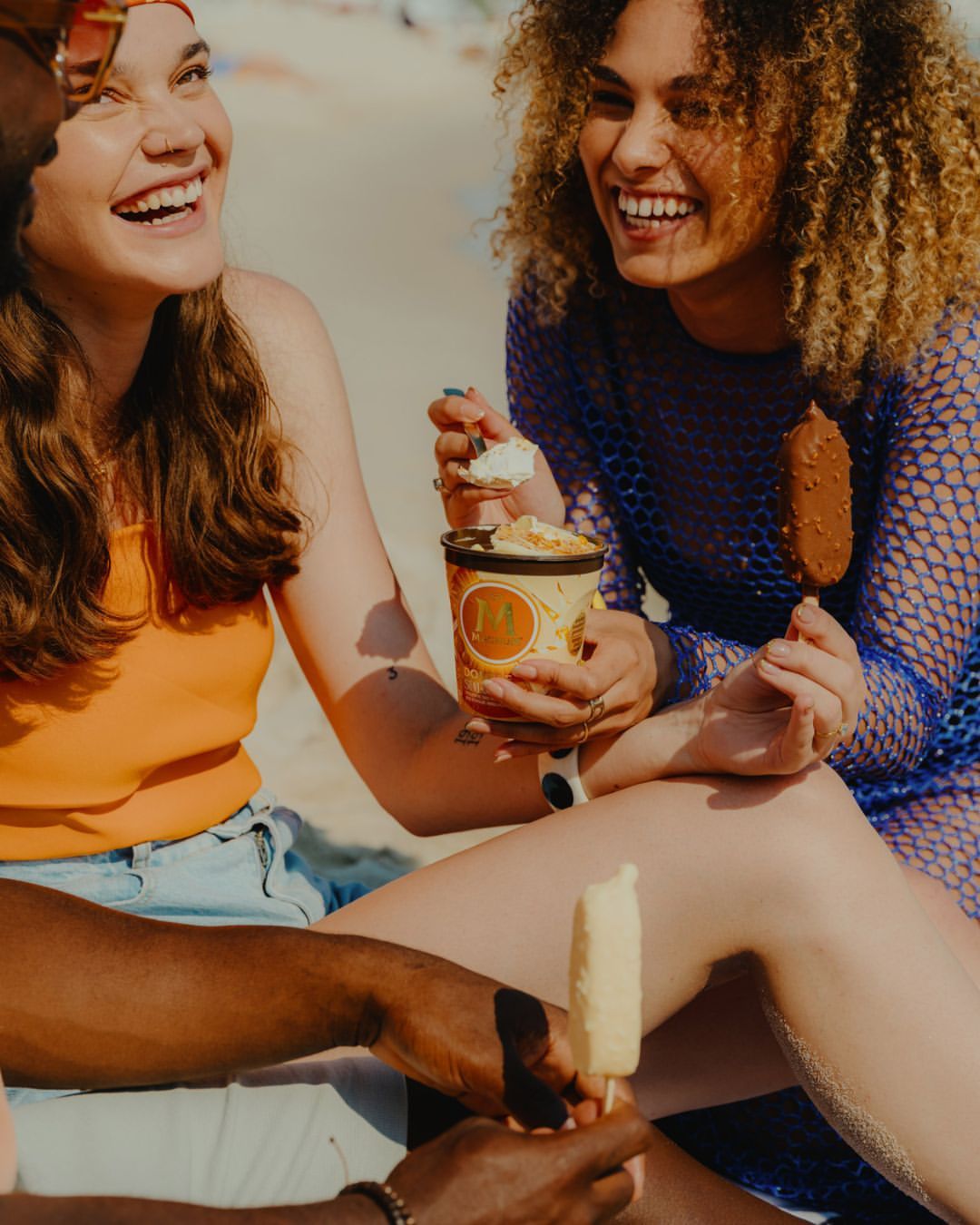 Group of friends laughing on a beach while eating ice cream, enjoying a sunny day with blue sky in a lifestyle, summertime setting.
