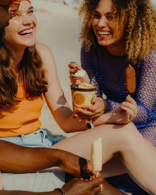 Group of friends laughing on a beach while eating ice cream, enjoying a sunny day with blue sky in a lifestyle, summertime setting.