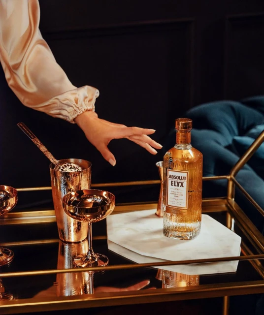 Elegant still life shot of Absolut Elyx bottle and copper barware on a bar cart with a hand reaching for the bar setup.