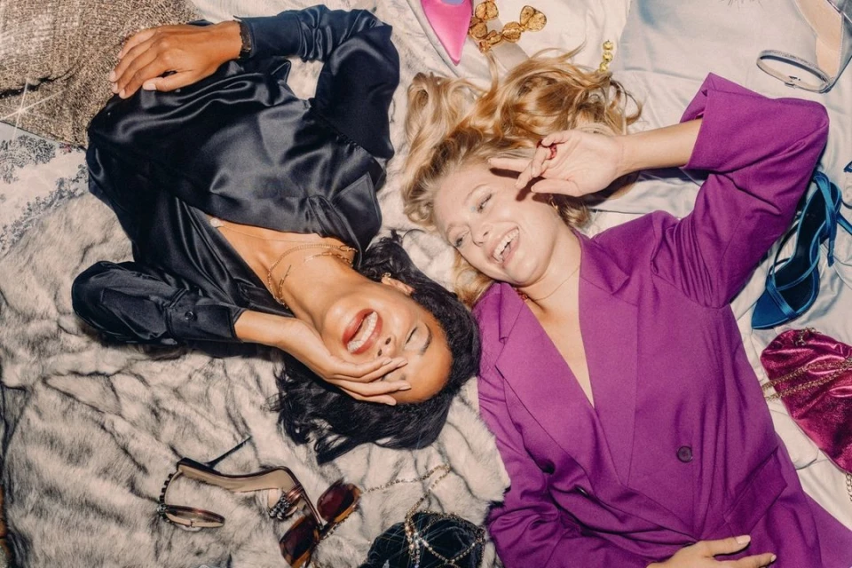 Two smiling women lying on a bed covered in faux fur, surrounded by shoes and purses. One woman wears a purple blazer and the other, a black satin top.