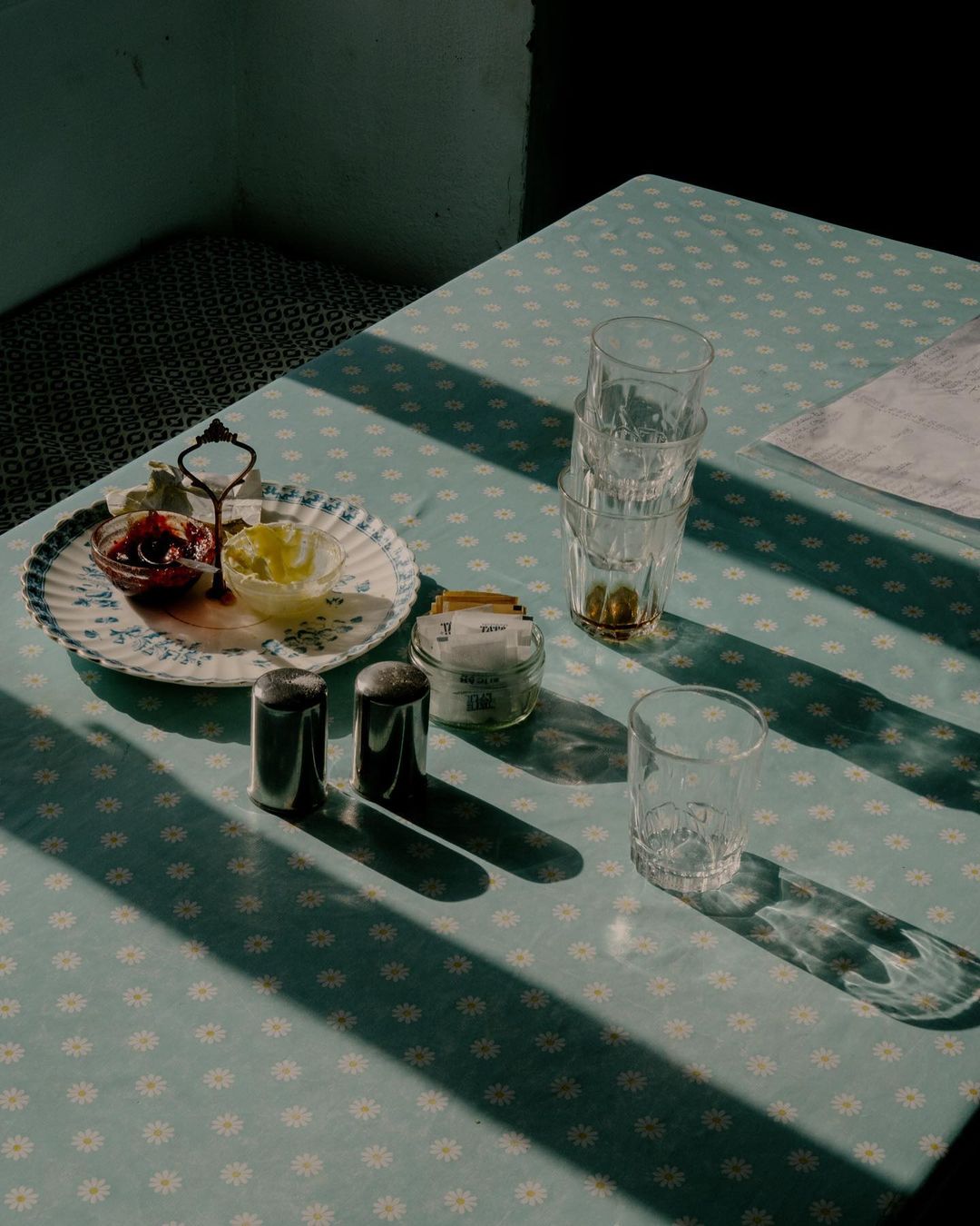 Overhead shot of a table with a patterned tablecloth, featuring a stack of drinking glasses, condiments, and a patterned plate with food.