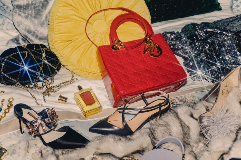 Still life of luxury fashion accessories including a red quilted handbag, jewelry, high heels, perfume bottle, and pillows on a bed.