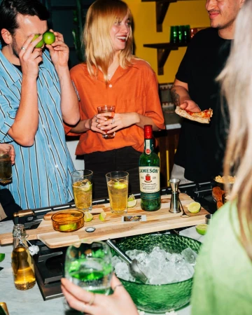 Group of friends at a casual indoor gathering with cocktails, a bottle of Jameson, pizza and ice. People are laughing and holding drinks.