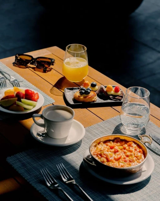 Overhead shot of a wooden table with fruit, pastries, a coffee cup, juice, silverware, and sunglasses for a bright, inviting breakfast.