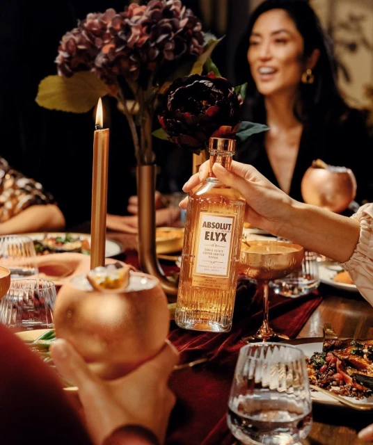 Absolut Elyx vodka bottle on a dinner table with drinks, food, flowers, and people in the background, creating an elegant, festive atmosphere.
