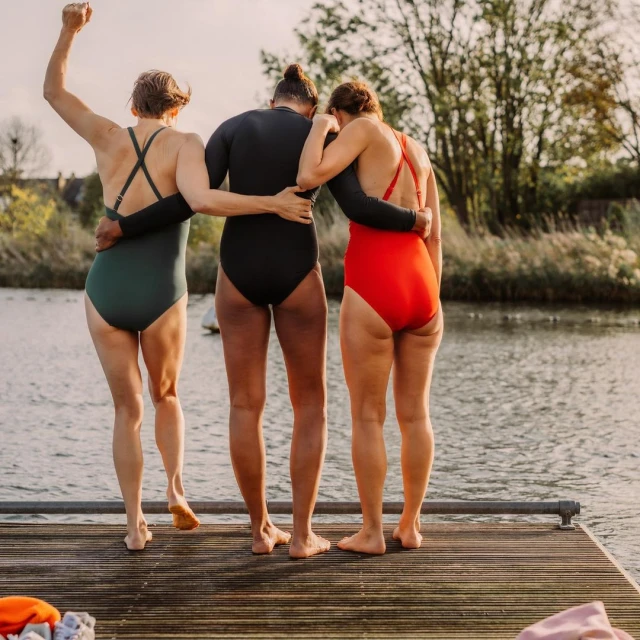 Three women in swimsuits stand on a wooden dock near a lake on a sunny day, one with her arm raised in the air in victory.