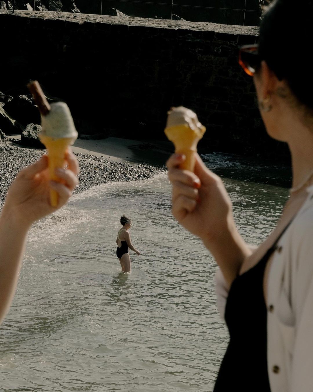 Two people hold ice cream cones on a beach, with a bather visible in the water on a sunny day, capturing a summertime mood.