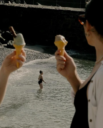 Two people hold ice cream cones on a beach, with a bather visible in the water on a sunny day, capturing a summertime mood.