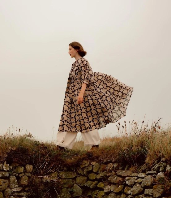 Adult woman in a patterned dress walks on a stone wall outdoors with a cloudy sky, showcasing fashion, lifestyle and nature.