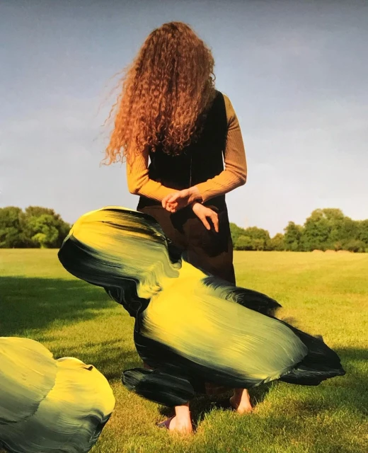 Back view of a red-haired woman in a field, wearing a yellow top and a dark vest, standing near abstract black and yellow objects.