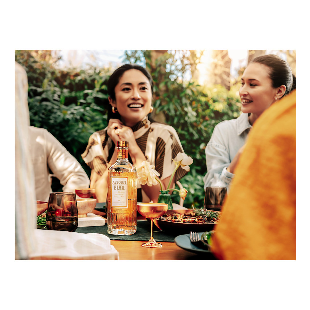 A group of young adults sit at a table on a patio with a bottle of Absolut Elyx vodka, food, and drinks, surrounded by greenery.