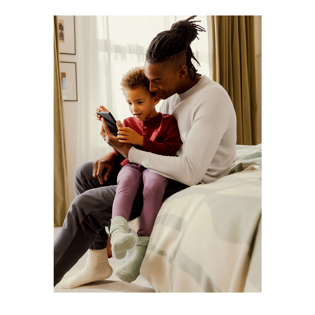 Dad and son enjoy mobile device together indoors. Father sits on bed with young child playing on phone; casual, intimate family time.