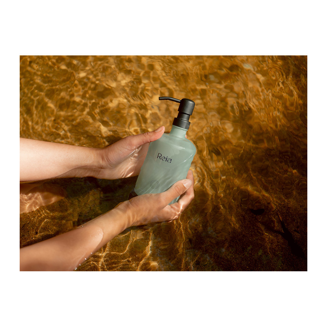Hands hold a light green bottle with black pump in shallow, clear water. The bottle has a white 'Reia' logo on its side.