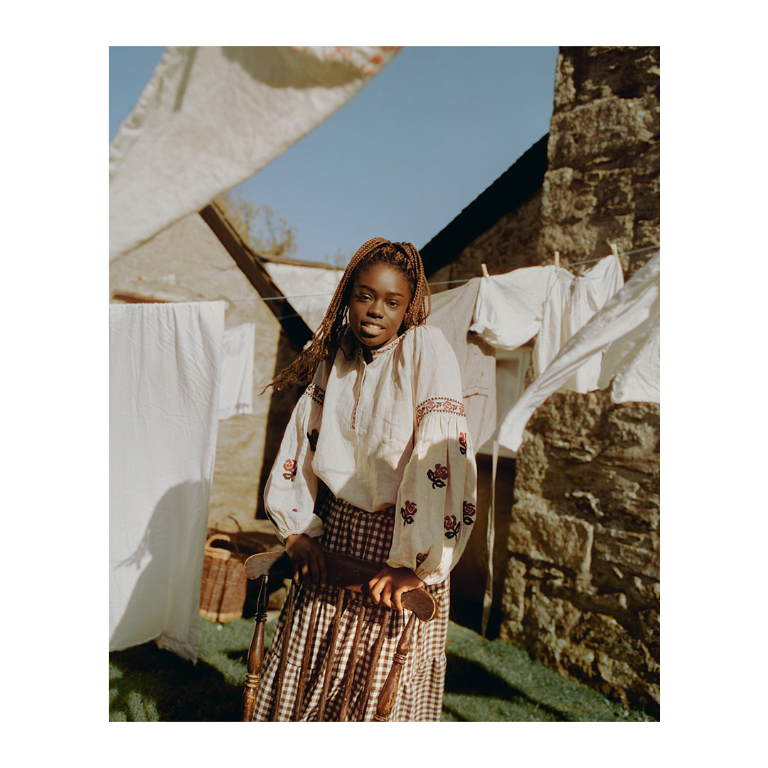 A young Black woman in an embroidered blouse and skirt poses outdoors on a sunny day with a clothesline and stone building behind her.