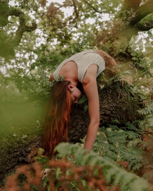 A woman with red hair rests in a tree surrounded by green leaves and branches in a serene outdoor setting, captured in a medium shot.