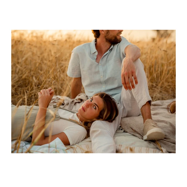 A couple lounges in a wheat field. The fair-skinned woman lies with her head resting in the man's lap as he sits behind her.