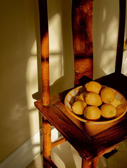 Still life of lemons in a shallow bowl sitting on a vintage wooden chair, illuminated by soft, warm window light.